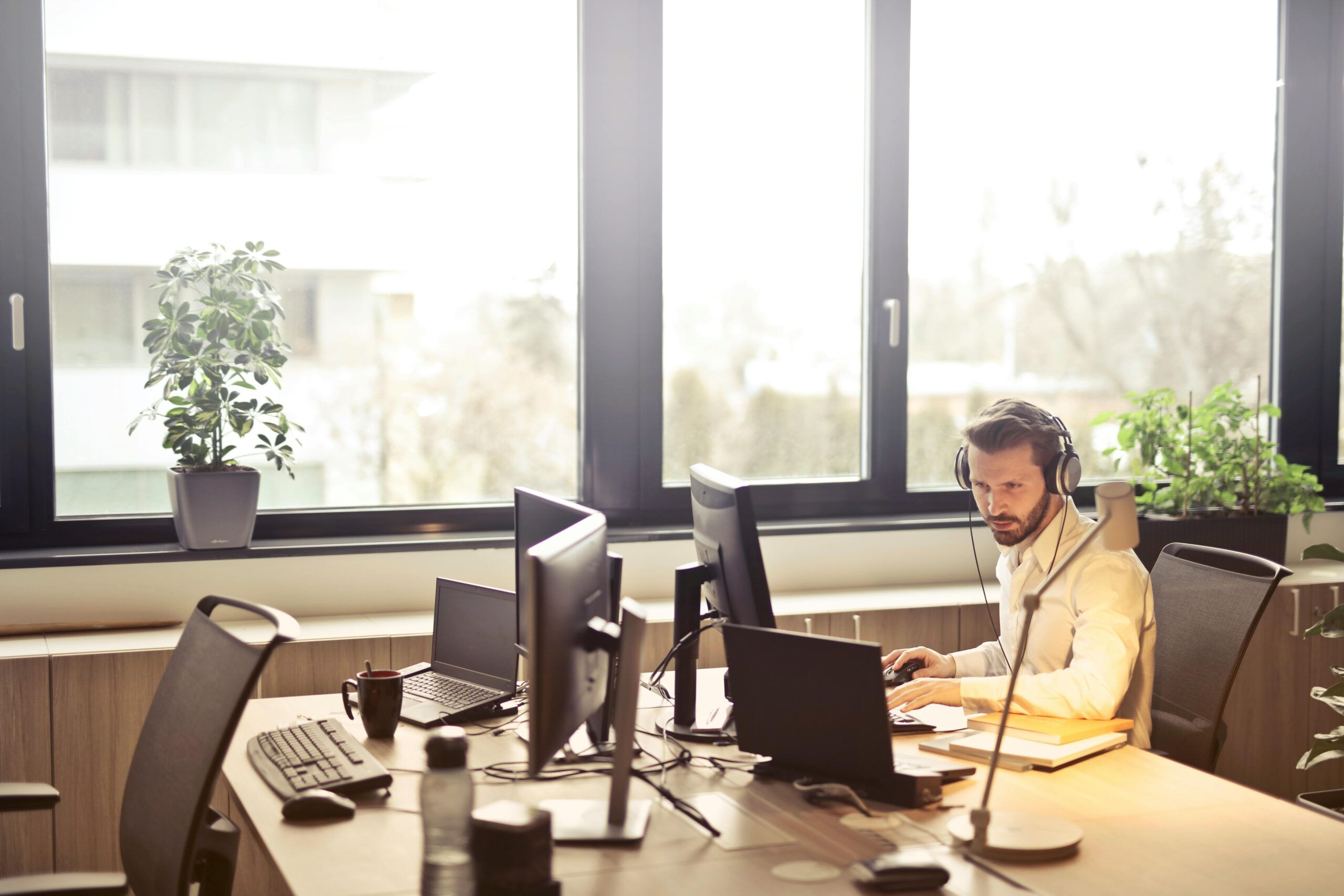 Chi siamo A businessman sits at a desk using multiple computers and a headset in a well-lit modern office.
